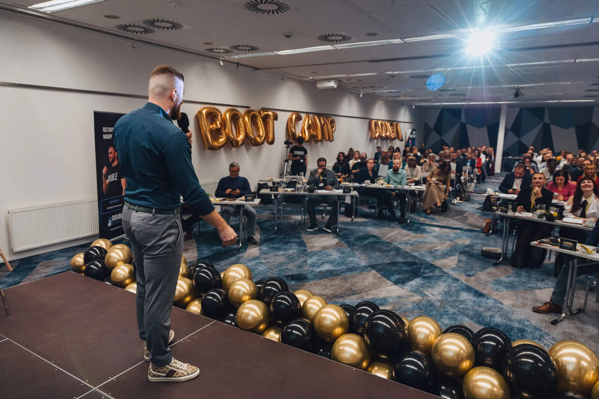 Řečník na pódiu mluví k plnému sálu, v pozadí zlaté balónkové písmo Boot Camp.