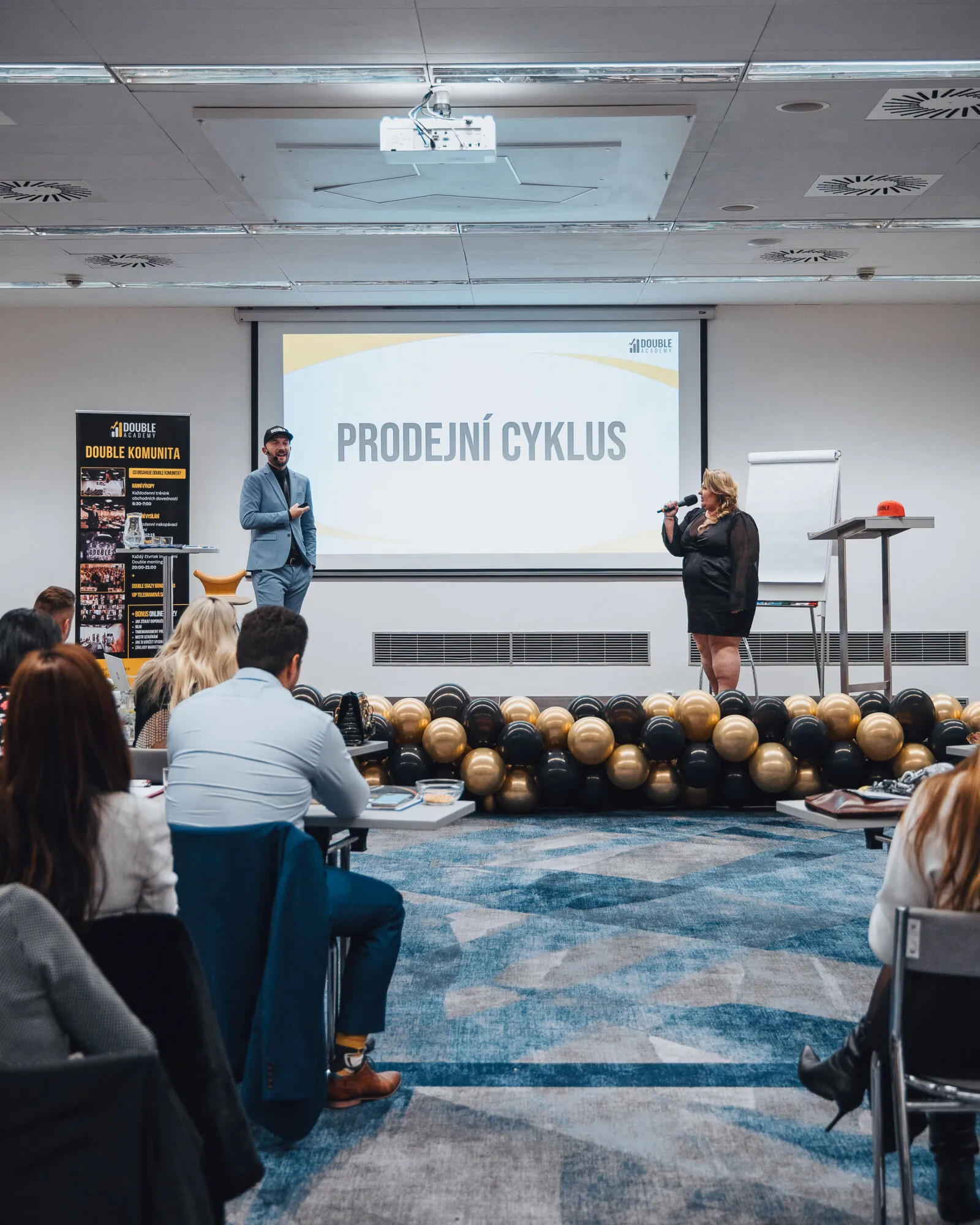 Dva řečníci na pódiu před prezentací s tématem Prodejní cyklus a publikem v sále.