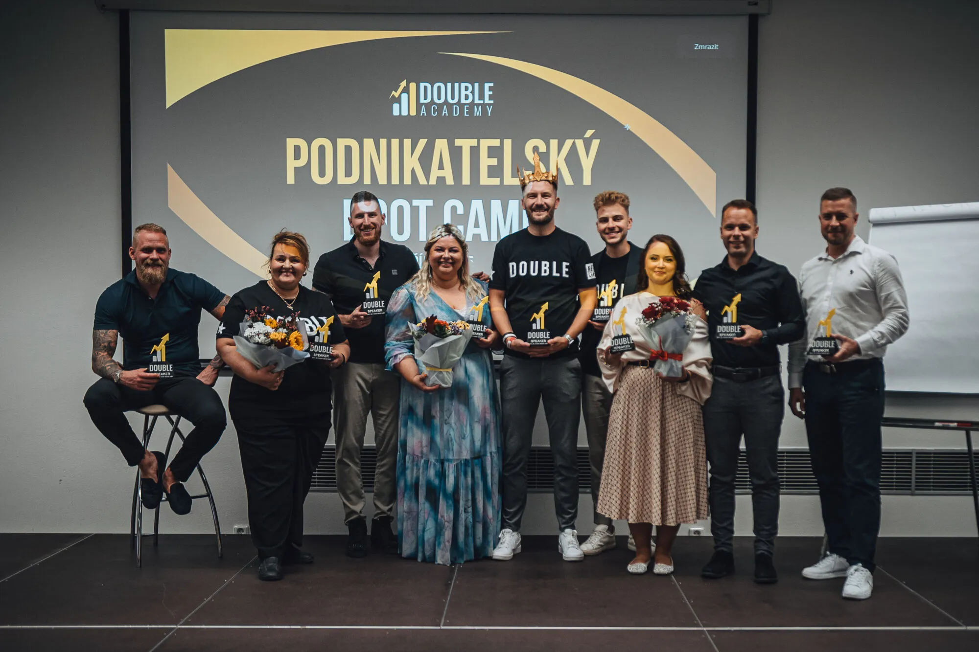 Skupina lidí s trofejemi a kyticemi pózuje před projekcí Podnikatelský Boot Camp Double Academy.
