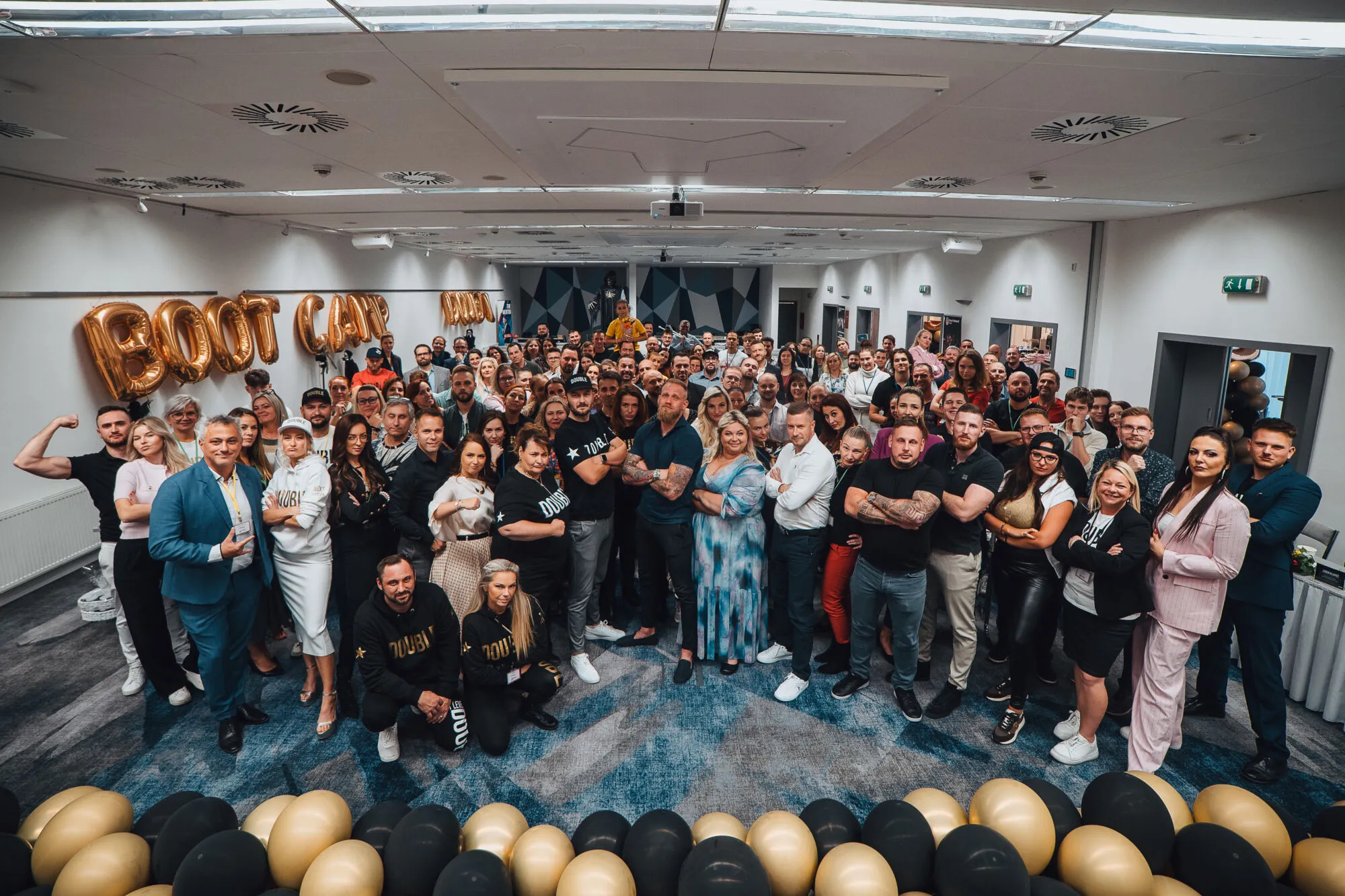 Skupinové foto velkého týmu a účastníků akce v sále se zlatými balónky s nápisem Boot Camp.